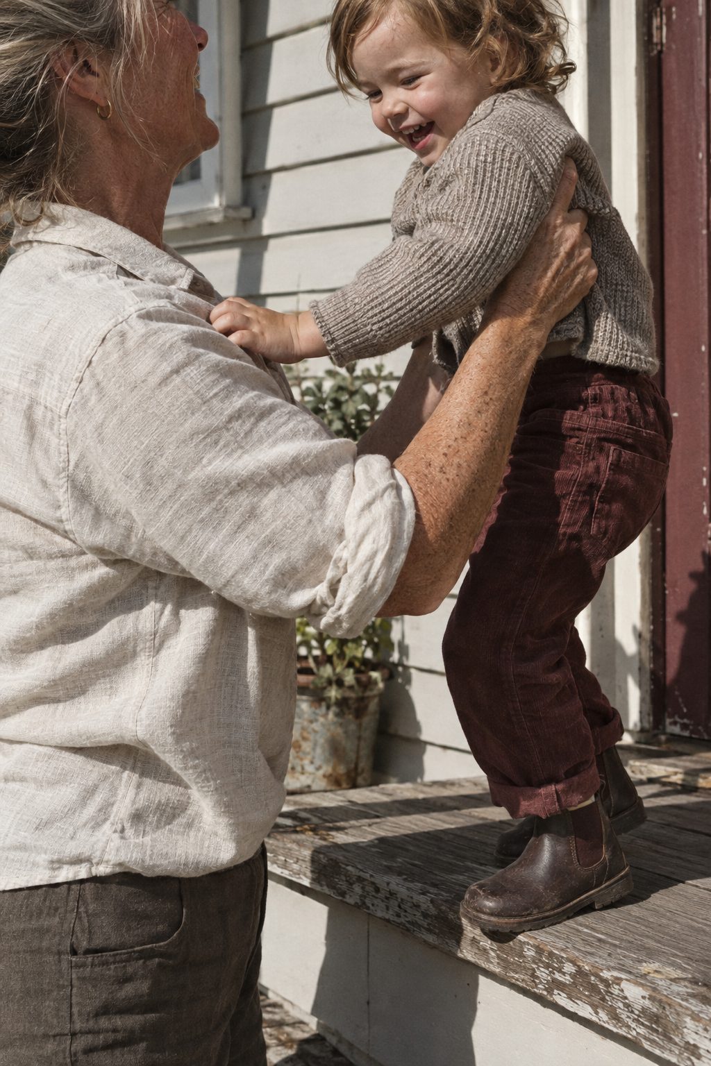 Forearms of a woman in her late fifties lifting a toddler onto a weathered timber back step in hard afternoon light